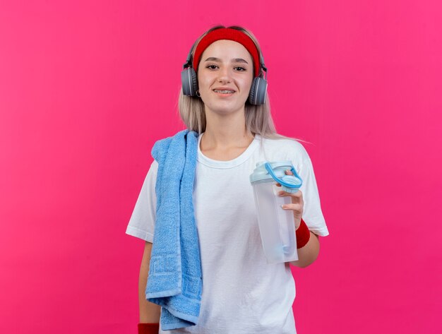 Улыбающаяся молодая спортивная женщина с подтяжками на наушниках, с повязкой на голову и браслетами держит бутылку с водой и полотенце на плече, изолированном на розовой стене