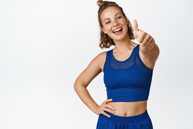 Smiling young spportswoman shows thumbs up. Motivated fitness woman like something, giving compliment, recommending sport gym, white background