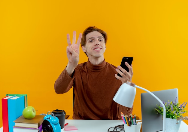 Улыбающийся молодой студент мальчик сидит за столом со школьными инструментами, держит телефон и показывает три изолированные на желтой стене