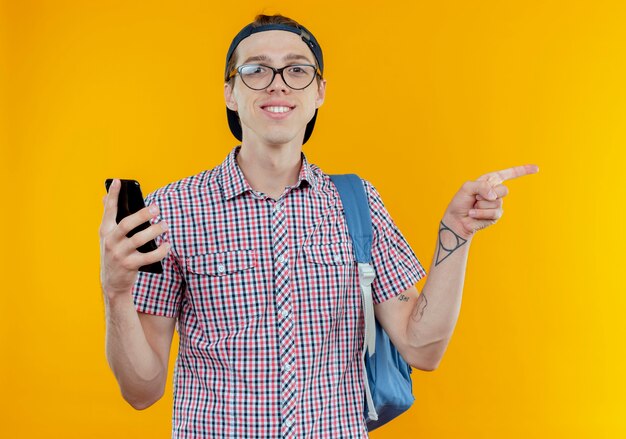 Smiling young student boy wearing back bag and glasses and cap holding phone and points at side on white