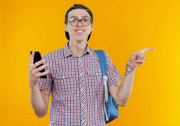 Smiling young student boy wearing back bag and glasses and cap holding phone and points at side on white