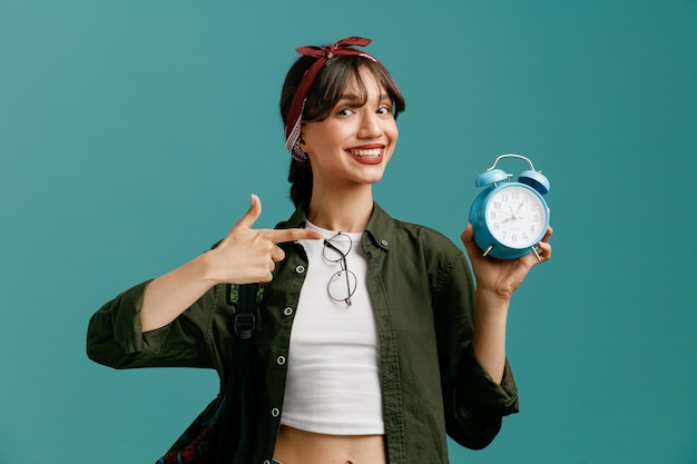 Foto gratuita sorridente giovane studentessa che indossa bandana e zaino guardando la fotocamera che mostra la sveglia che punta ad essa isolata su sfondo blu