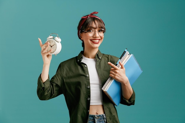 Smiling young student girl wearing bandana glasses holding large note pads with pen looking at camera showing alarm clock pointing at clock isolated on blue background