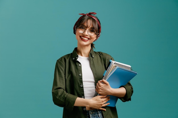 Smiling young student girl wearing bandana glasses holding large note pads with pen with both hands looking at camera isolated on blue background