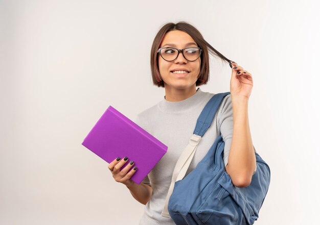 Smiling young student girl wearing glasses and back bag holding book and her hair looking at side isolated on white wall