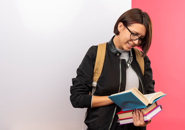 웃는 젊은 학생 소녀 안경을 쓰고 다시 가방을 들고 분홍색 벽에 고립 된 흰 벽 앞에 서있는 책을보고