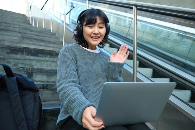 웃고 있는 어린 학생은 헤드폰을 끼고 거리 계단에 앉아 그녀의 노트북에서 손을 흔들며 t를 연결합니다.