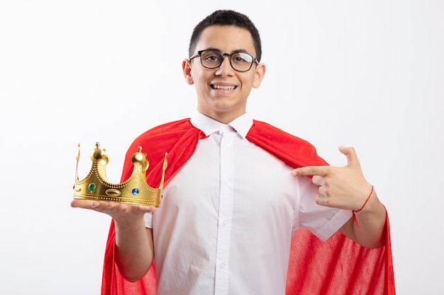 Sorridente giovane supereroe ragazzo in mantello rosso con gli occhiali tenendo e indicando la corona che guarda l'obbiettivo isolato su priorità bassa bianca