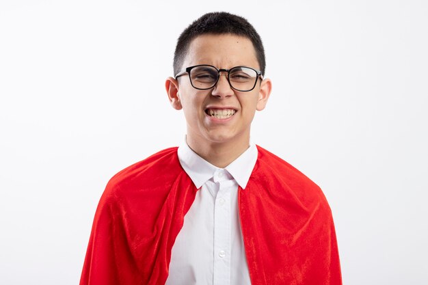 Smiling young superhero boy in red cape wearing glasses looking at camera isolated on white background
