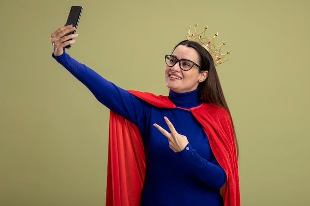 Smiling young superhero girl wearing glasses and crown showing peace gesture take a selfie isolated on olive green background