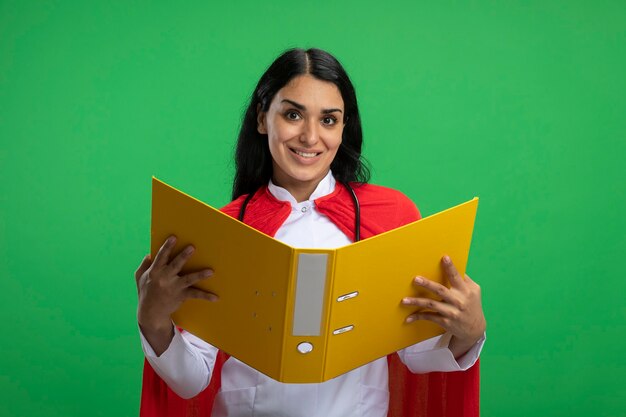 Улыбающаяся молодая девушка супергероя в медицинском халате со стетоскопом, держащая папку, изолированную на зеленом