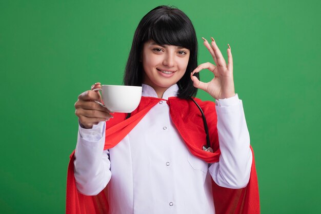 Smiling young superhero girl wearing stethoscope with medical robe and cloak holding cup of tea showing okay gesture isolated on green wall