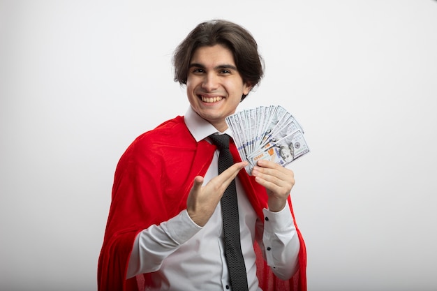 Smiling young superhero guy looking at camera wearing tie holding and points at cash isolated on white