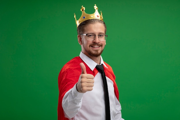 Бесплатное фото Улыбающийся молодой супергерой в короне показывает большой палец вверх изолирован на зеленом фоне
