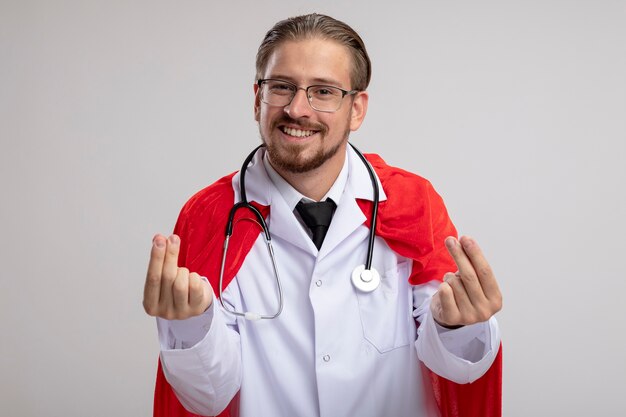 Smiling young superhero guy wearing medical robe with stethoscope and glasses showing tip gesture isolated on white background