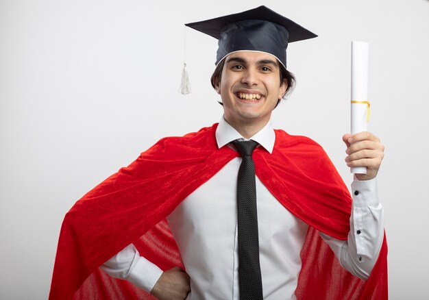 Smiling young superhero guy wearing tie and graduate hat holding diploma and putting hand on hip