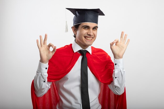 ネクタイと白い背景で隔離の大丈夫なジェスチャーを示す卒業帽子を身に着けている若いスーパーヒーローの男を笑顔