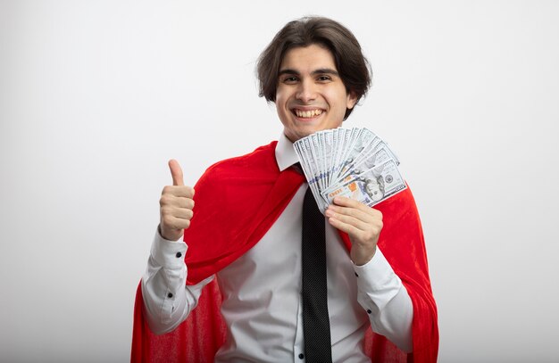 Smiling young superhero guy wearing tie holding cash and showing thumb up isolated on white