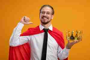 Free photo smiling young superhero guy wearing tie holding crown and showing thumb up isolated on orange background