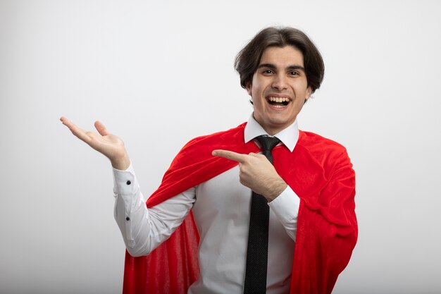 Smiling young superhero guy wearing tie pretending holding and points at something isolated on white