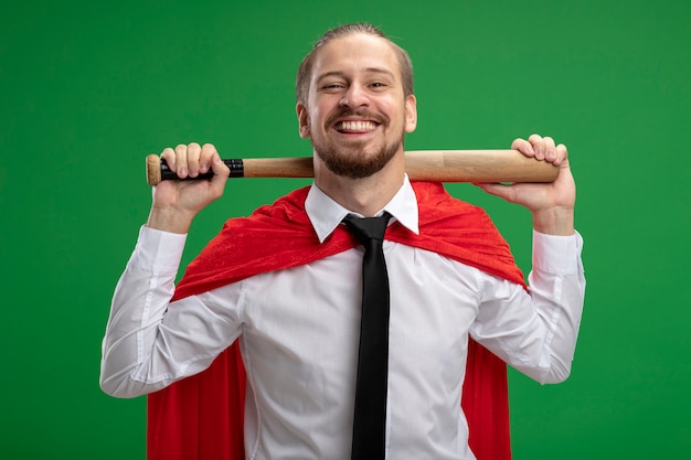 Smiling young superhero guy wearing tie putting baseball bat on neck isolated on green