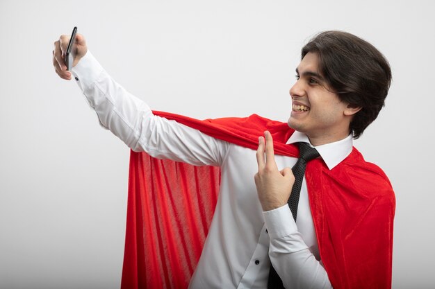 Smiling young superhero guy wearing tie take a selfie and showing peace gesture isolated on white background