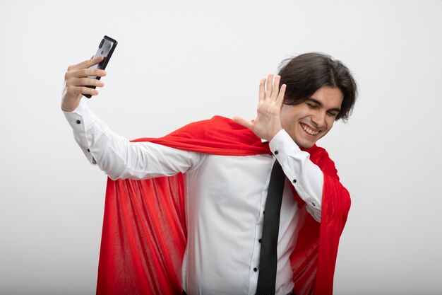 Smiling young superhero guy with closed eyes wearing tie take a selfie and covered face with hand isolated on white background