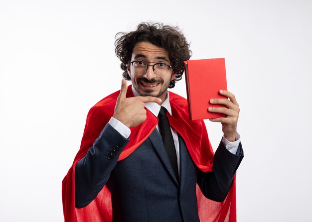 Улыбающийся молодой супергерой в оптических очках в костюме с красным плащом держит и указывает на книгу, изолированную на белой стене