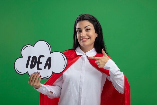 Smiling young superwoman holding idea bubble and pointing her index finger to it looking at front isolated on green wall