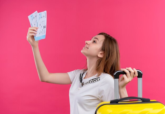 Smiling young traveler girl holding airplane tickets and suitcase looking up on isolated pink wall