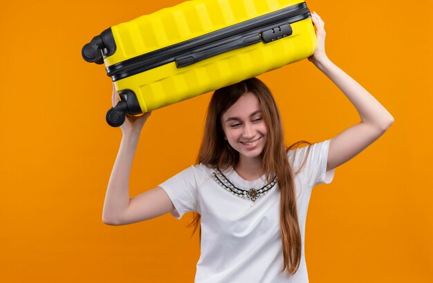 Smiling young traveler girl putting suitcase on head on isolated orange wall