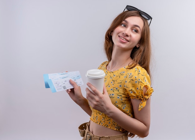 Улыбающаяся молодая путешественница в солнцезащитных очках на голове держит билеты на самолет и пластиковую кофейную чашку на изолированной белой стене