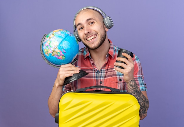 복사 공간이 있는 보라색 벽에 격리된 가방 뒤에 서 있는 글로브와 종이 컵을 들고 헤드폰을 끼고 웃고 있는 젊은 여행자