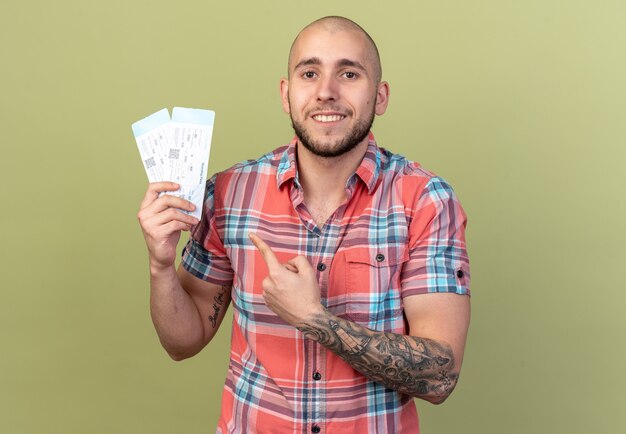 smiling young traveler man holding and pointing at air tickets isolated on olive green wall with copy space