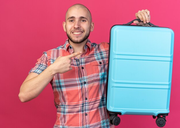 улыбающийся молодой путешественник, держащий и указывающий на чемодан, изолированный на розовой стене с копией пространства
