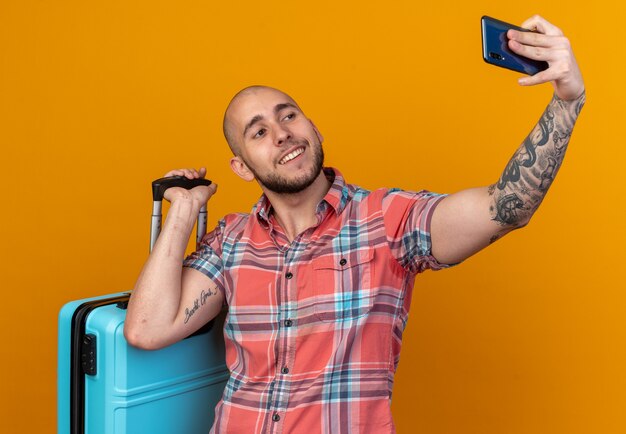 smiling young traveler man holding suitcase and taking selfie on phone isolated on orange wall with copy space