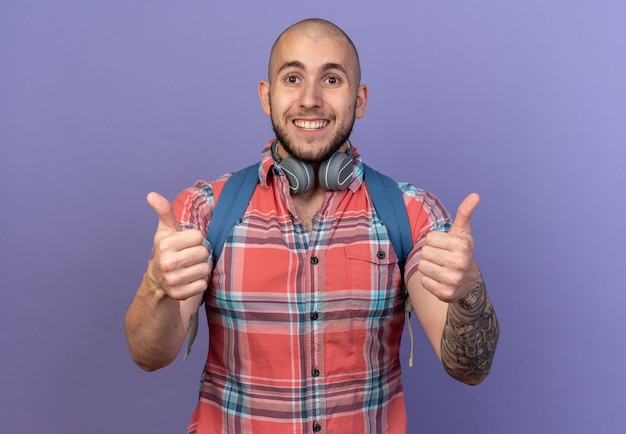 Smiling young traveler man with headphones around his neck and with backpack thumbing up isolated on purple wall with copy space