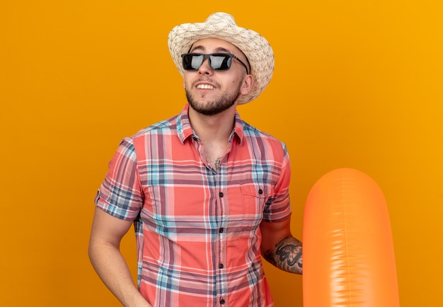 smiling young traveler man with straw beach hat in sun glasses holding swim ring looking at side isolated on orange wall with copy space