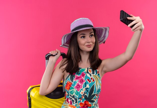 Smiling young traveler woman doing selfie holding suitcase on isolated pink wall