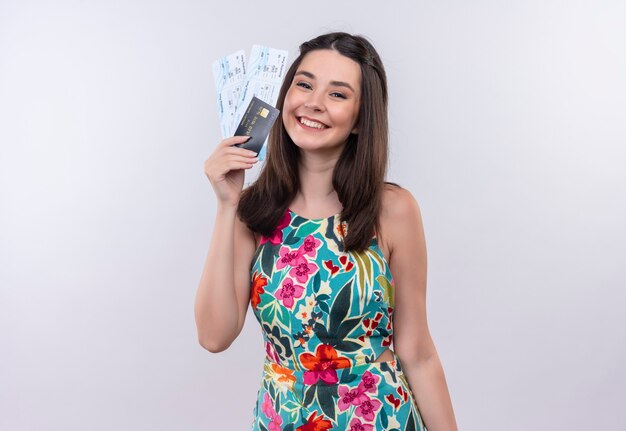 Smiling young traveler woman wearing multicolor dress holding a tickets and card on white wall