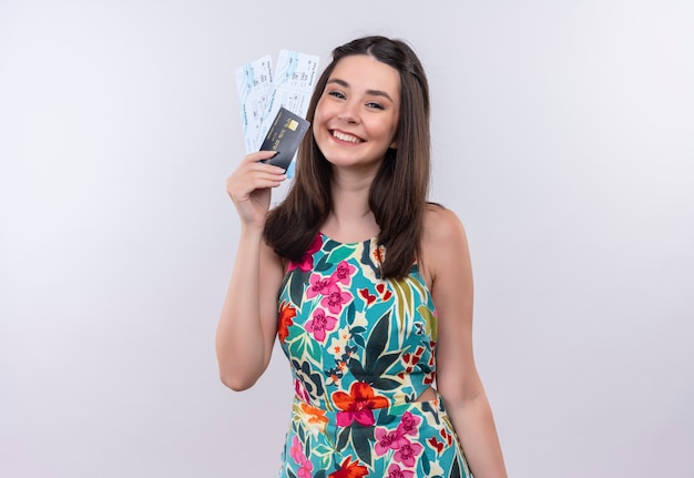 Улыбающаяся молодая женщина-путешественница в разноцветном платье держит билеты и открытку на белой стене