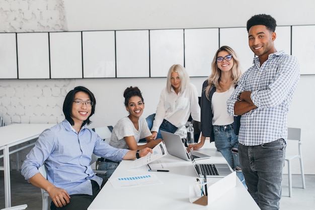 Foto gratuita sorridenti giovani sviluppatori web in posa intorno al tavolo con i laptop su di esso. ritratto dell'interno della studentessa asiatica con capelli neri che trascorre del tempo con gli amici all'università.