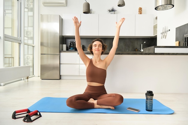 Free photo smiling young woman in activewear sitting on mat at home raising arms up while meditating practice y