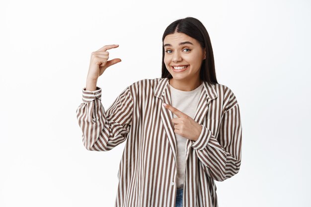 Smiling young woman awkwardly pointing at small size gesture, showing little thing, tiny item in hand, standing against white wall