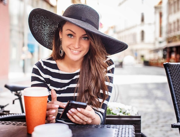 Улыбающаяся молодая женщина в черной рубашке с белыми полосками и большой черной летней шляпе сидит за столом в летнем кафе.