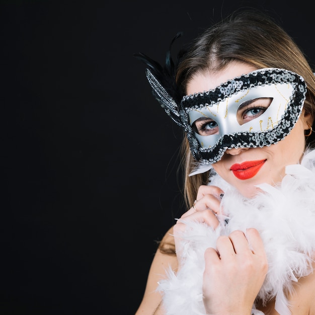 Улыбающаяся молодая женщина в карнавальной маске держит боа на черном фоне