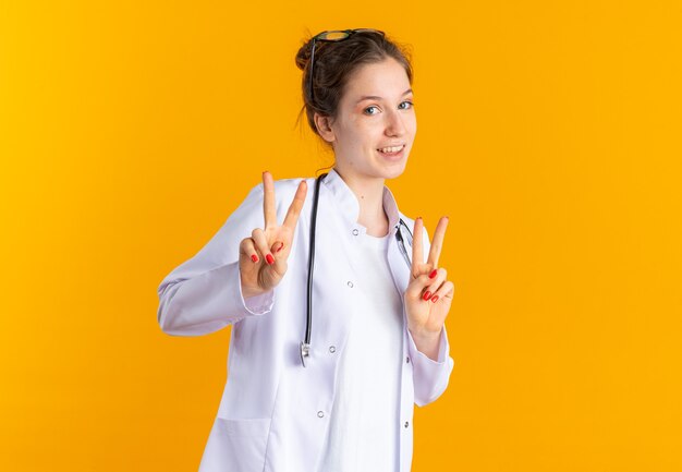 두 손으로 승리 기호를 몸짓으로 청진기를 들고 의사 제복을 입은 웃는 젊은 여성