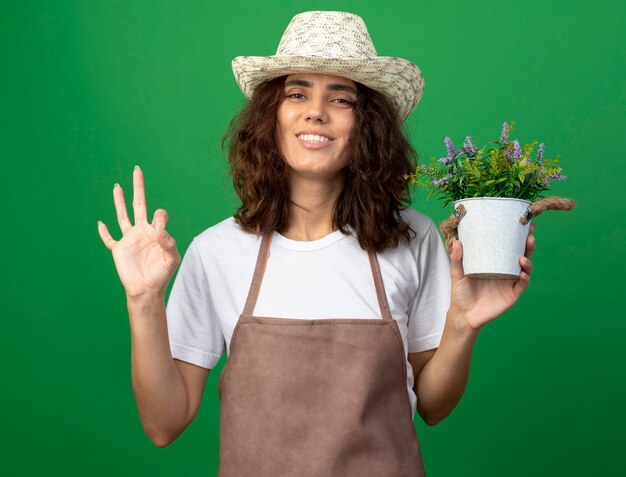 Smiling young woman gardener in uniform wearing gardening hat holding flower in flowerpot and showing okay gesture isolated on green