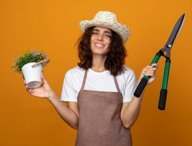 Foto gratuita giardiniere sorridente della giovane donna in uniforme che indossa il cappello di giardinaggio che tiene il fiore in vaso di fiori con i clippers isolati sull'arancio