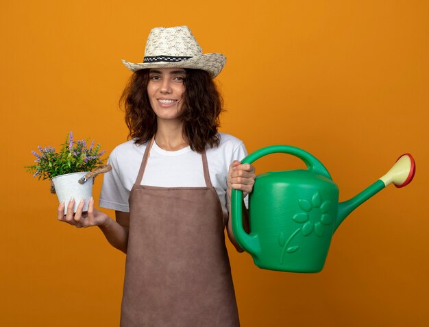 Smiling young woman gardener in uniform wearing gardening hat holding watering can with flower in flowerpot isolated on orange